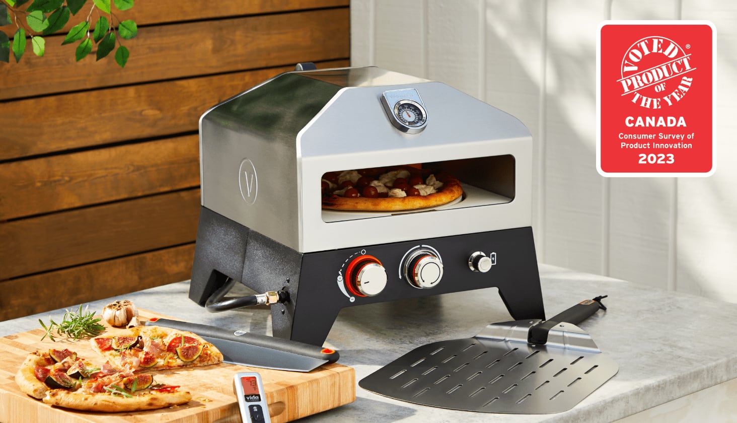 A Vida by PADERNO Gas Pizza Oven cooking pizza on a table, with a cooked pizza on a cutting board next to it.