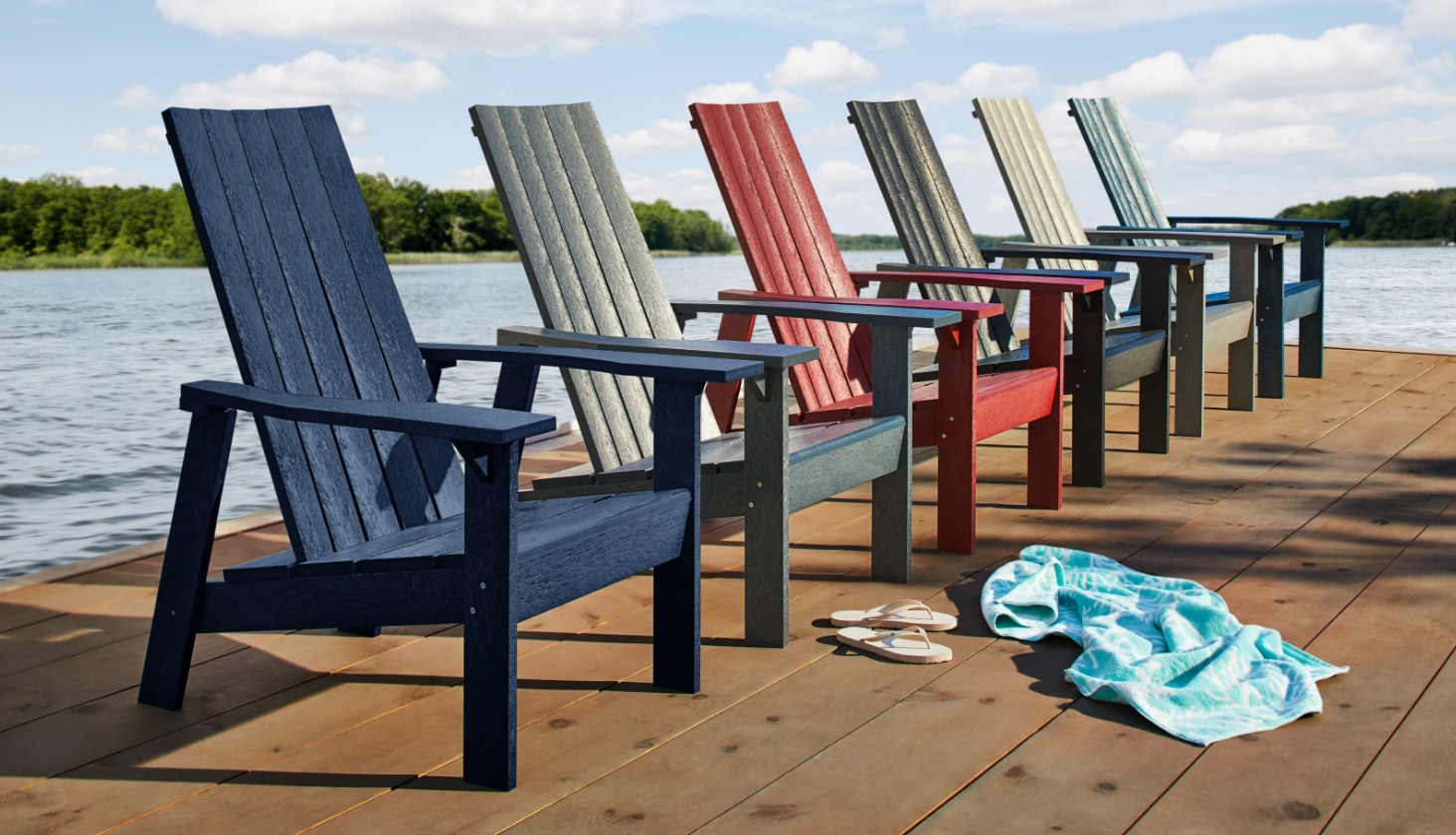A row of 6 CANVAS Arrowhead Muskoka Chairs lined up along a dock on a lake, with a pair of slippers and a towel laying on the dock in front of them.