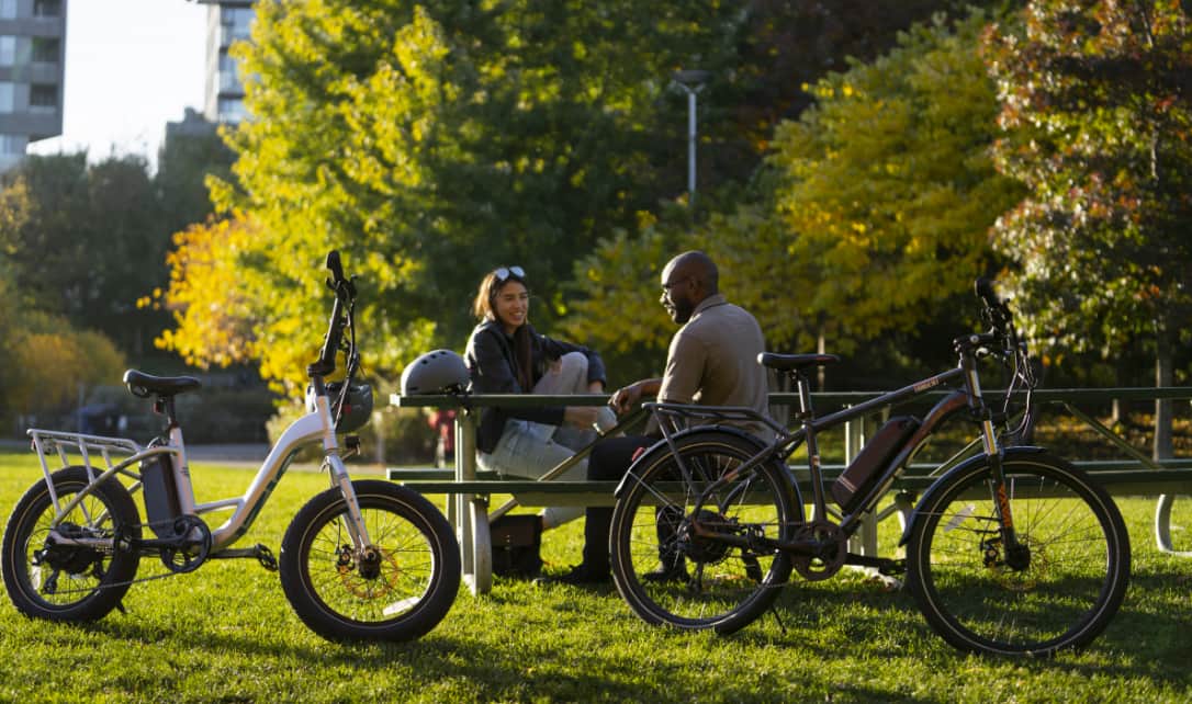 Two adults at a picnic bench with parked iZip e-bikes. 