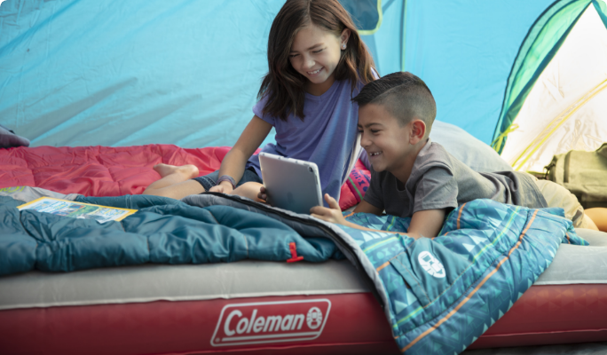 Two children sitting on an air mattress inside of a tent, playing on a tablet.