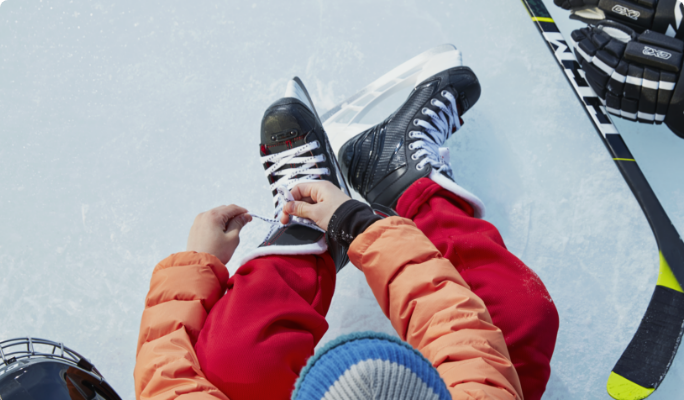 An overhead shot of someone sitting on an ice rink while lacing up their hockey skates with their helmet, hockey stick and gloves next to them.