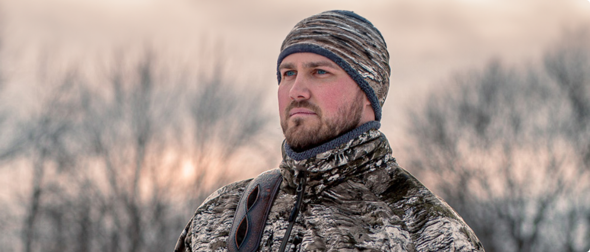 A man standing in front of trees in the winter at dusk, wearing a camouflage-patterned hat and jacket.