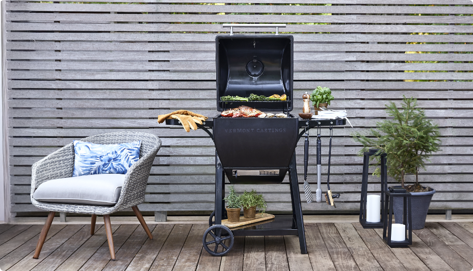 A Vermont Castings Pioneer™ Charcoal Kamado BBQ Grill next to plants and a single patio chair in front of a wooden fence on a deck.