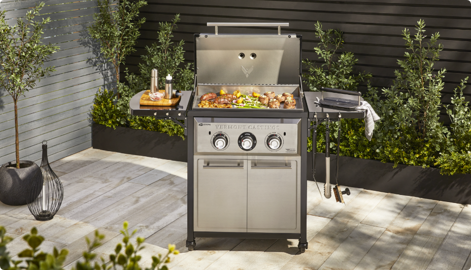 A Vermont Castings 2-Burner Propane Gas Griddle with TempASSURED Technology™ on a patio surrounded by greenery.