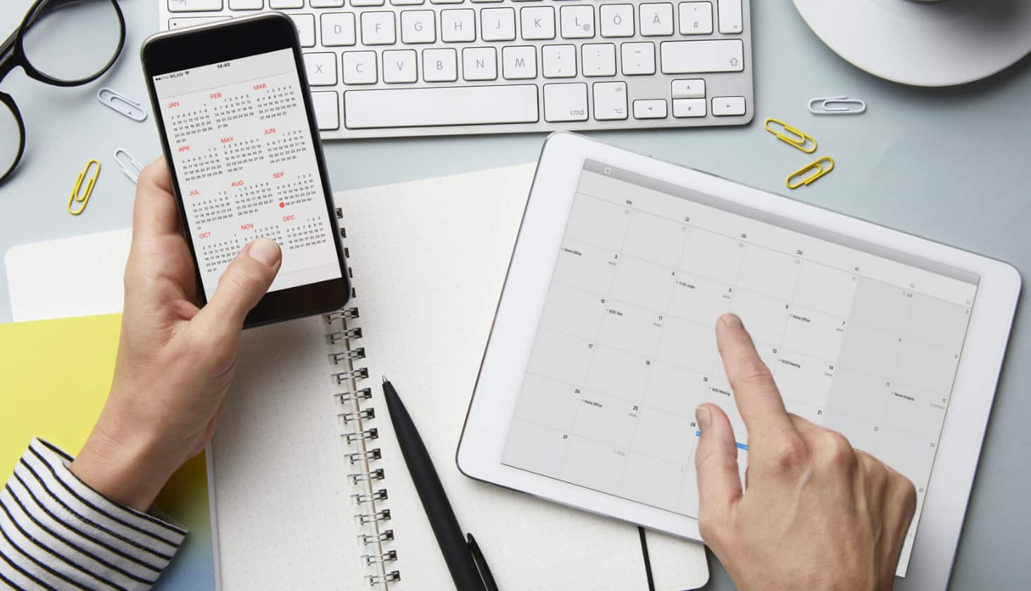 An overhead shot of hands holding a phone and pointing at a calendar on a tablet.