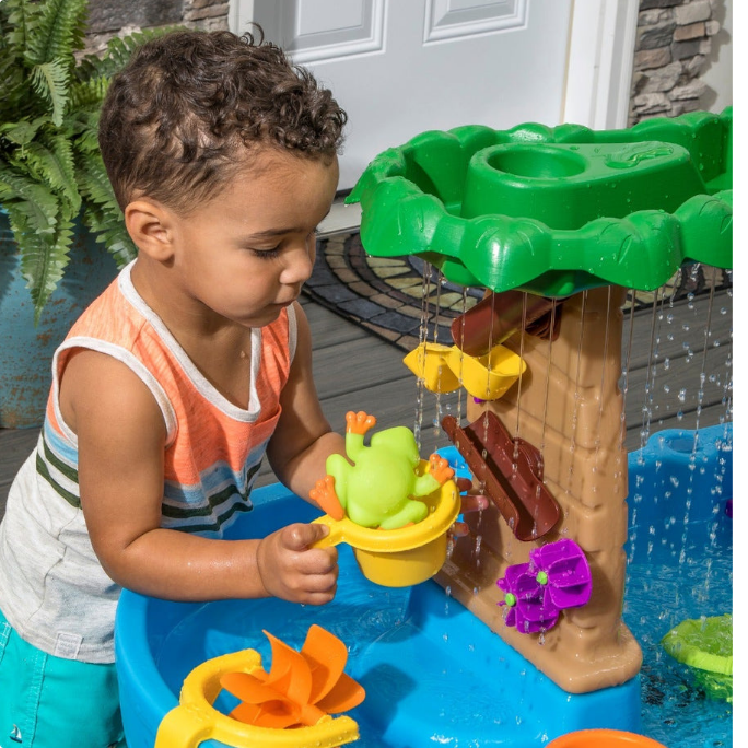 Un jeune enfant jouant avec une table d'activités aquatiques comprenant un arbre, des fleurs et un jouet grenouille. 