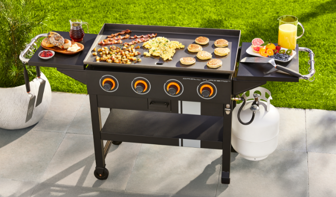 A griddle cooking up various foods with various cooked foods and accessories on the BBQ's side tables.