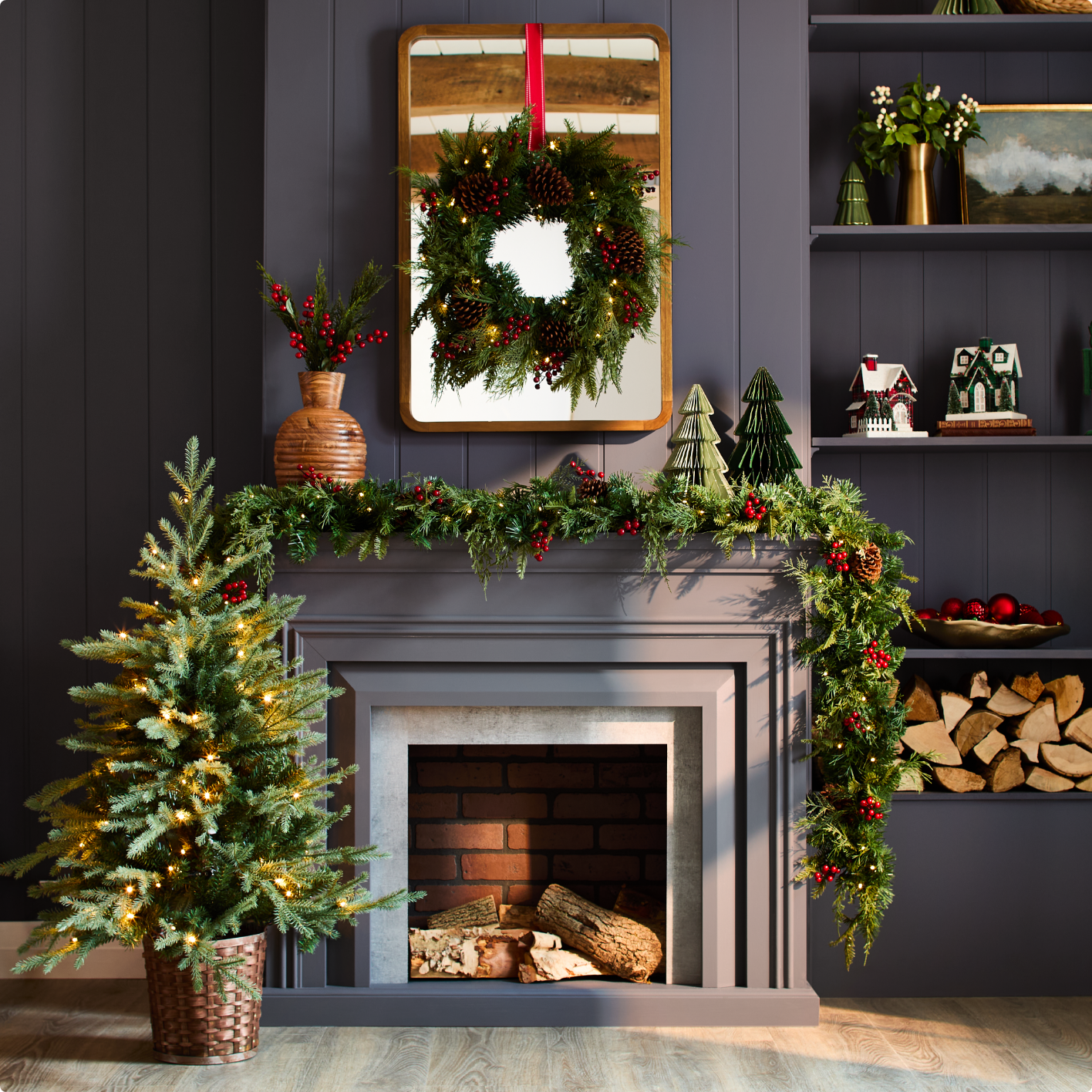 A festive fireplace with garland, a wreath on the mirror above, and a small Christmas tree next to it.
