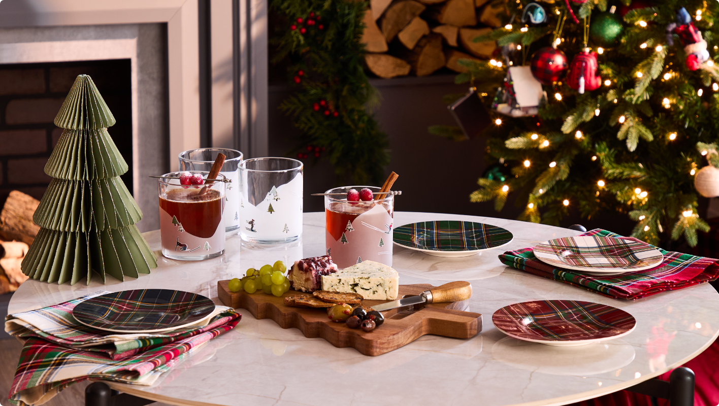 A festive table setting with plaid plates, drinks, a cheese board, and a green Christmas tree decoration.