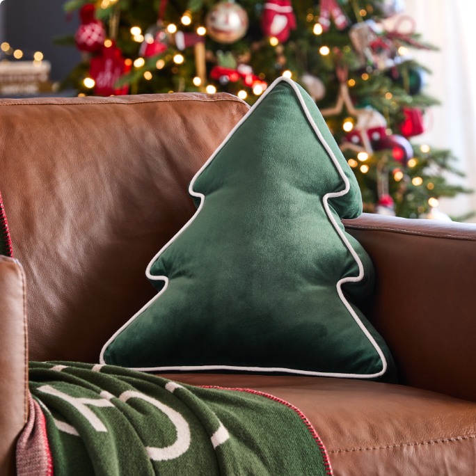 A green Christmas tree-shaped pillow and a festive blanket on a brown chair, with a decorated Christmas tree in the background.