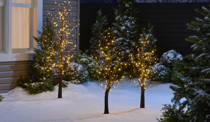 Des décorations de rennes scintillants CANVAS avec un arbre gemmé illuminé dans une cour enneigée. 