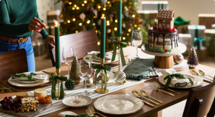 Table à manger festive garnie d'assiettes, de bougies vertes ornées de rubans, d'un gâteau et de décorations de Noël, près d'un sapin décoré.