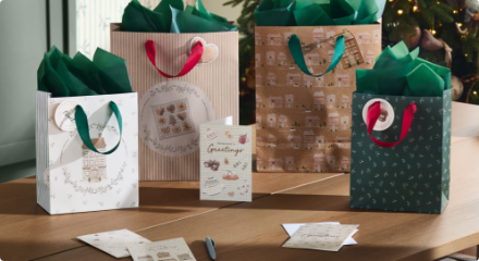 Quatre sacs cadeaux de Noël avec du papier de soie vert et une carte de vœux, posés sur un parquet en bois près d'un sapin de Noël décoré.