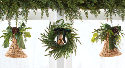 Petite couronne avec un ruban noir et une clochette, flanquée de deux clochettes en osier avec des boucles vertes, suspendues sous une guirlande devant une fenêtre.