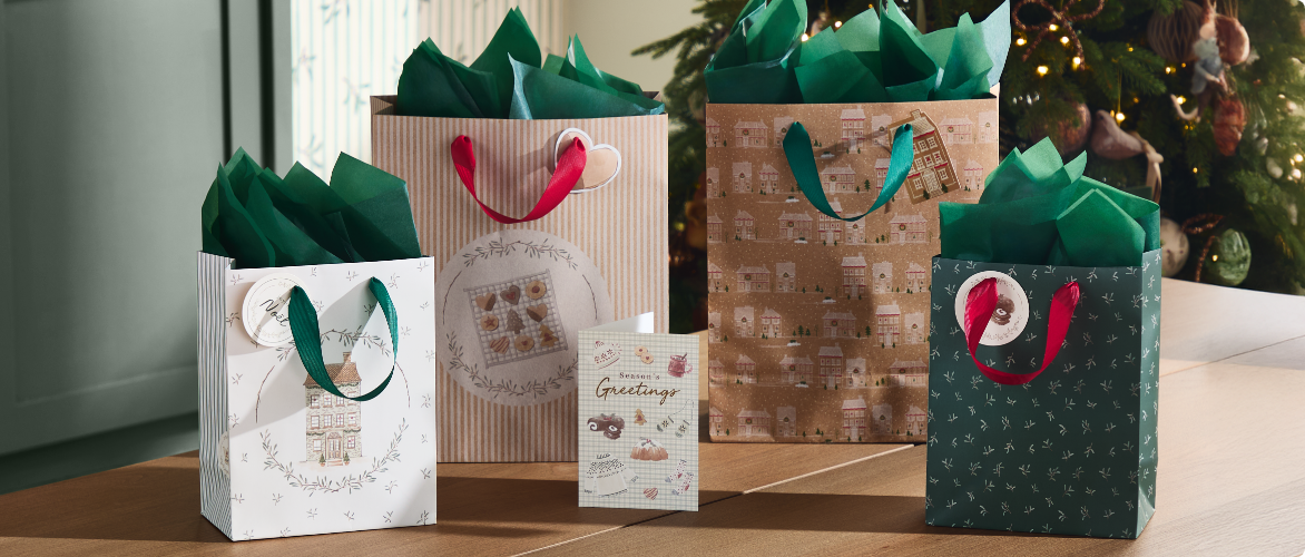 Quatre sacs cadeaux de Noël avec du papier de soie vert et une carte de vœux, posés sur un plancher en bois près d'un sapin de Noël décoré.
