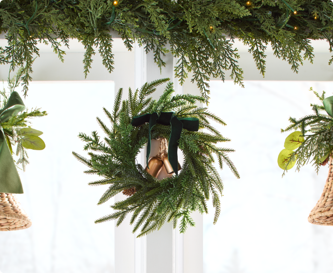 Une petite couronne ornée d'un ruban noir et d'une clochette, entourée de branches de verdure et de clochettes décoratives, devant une fenêtre.