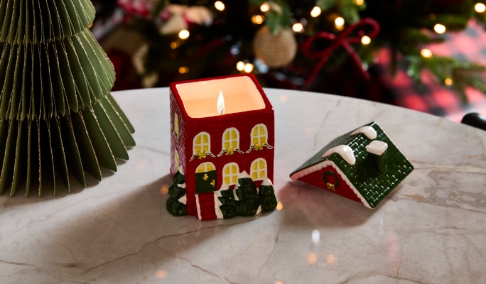 Une bougie allumée dans un porte-bougie en forme de maison avec un toit détaché est déposée sur une table, avec des décorations des Fêtes en arrière-plan.