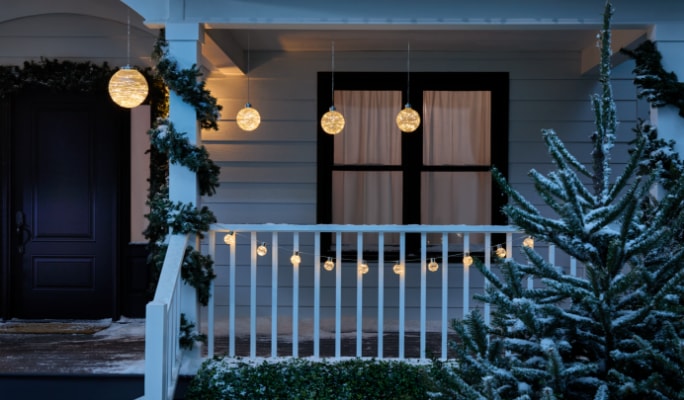Porche décoré de lumières suspendues, de guirlandes, et un arbre de Noël enneigé au premier plan.