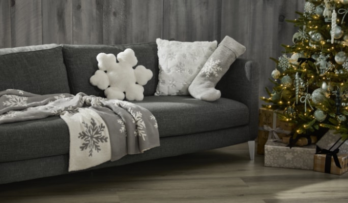 Canapé gris avec des coussins et des couvertures en forme de flocons de neige à côté d’un arbre de Noël décoré avec des cadeaux en dessous.