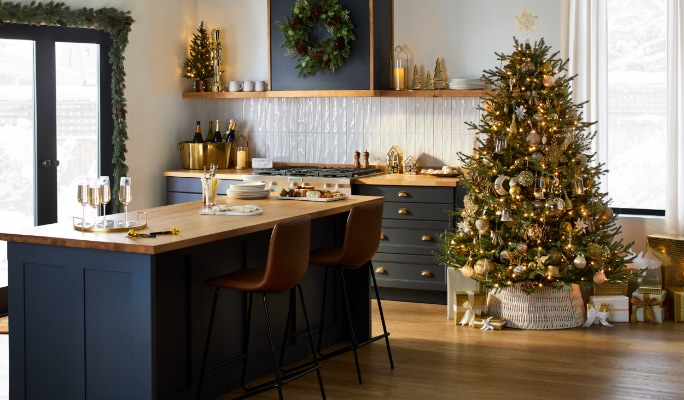 Cuisine décorée pour Noël avec un arbre, une couronne, des guirlandes et une table festive ornée d’accents dorés.