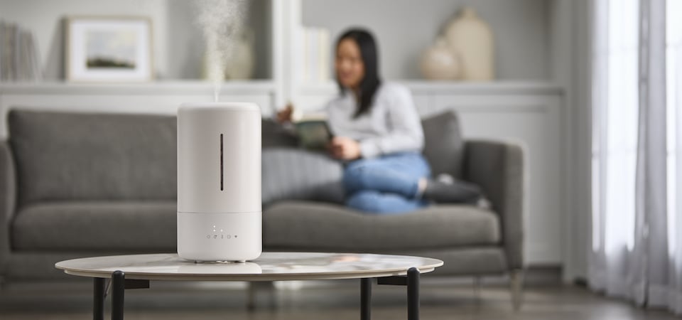 A person sits on a couch with a white air purifier on the table in the foreground.