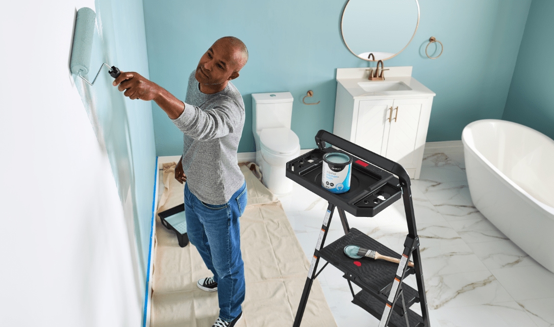 Une personne peignant les murs d'une salle de bain dotée d'un meuble-lavabo blanc, d'une baignoire autoportante et d'un beau décor.