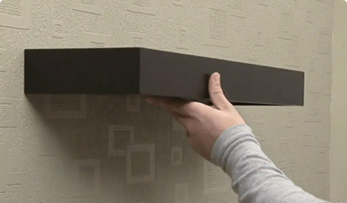 A hand holds a brown floating shelf against a wall.