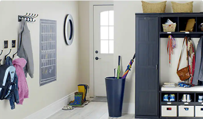 A home entryway with wall mirror, wall hooks with coats, umbrellas in a large bin, and  a coat and shoe storage unit in black.