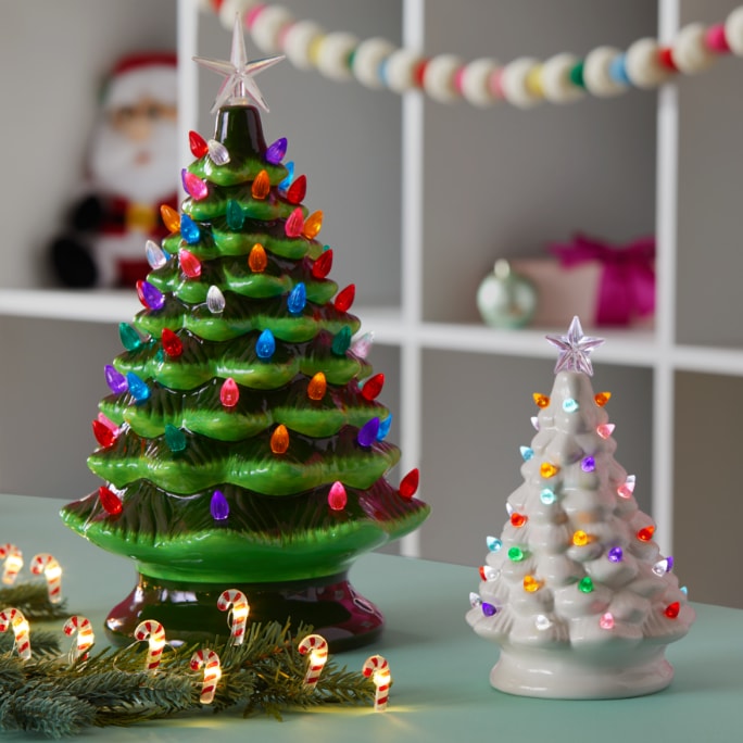 Two ceramic Christmas trees, one green and one white, are decorated with colorful lights and displayed on a table. 