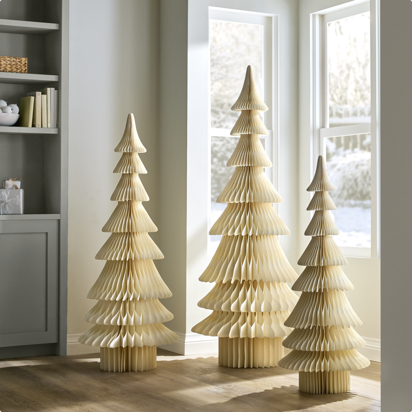 Three white Christmas trees made from paper,  in varying heights, in a bright room with large windows.