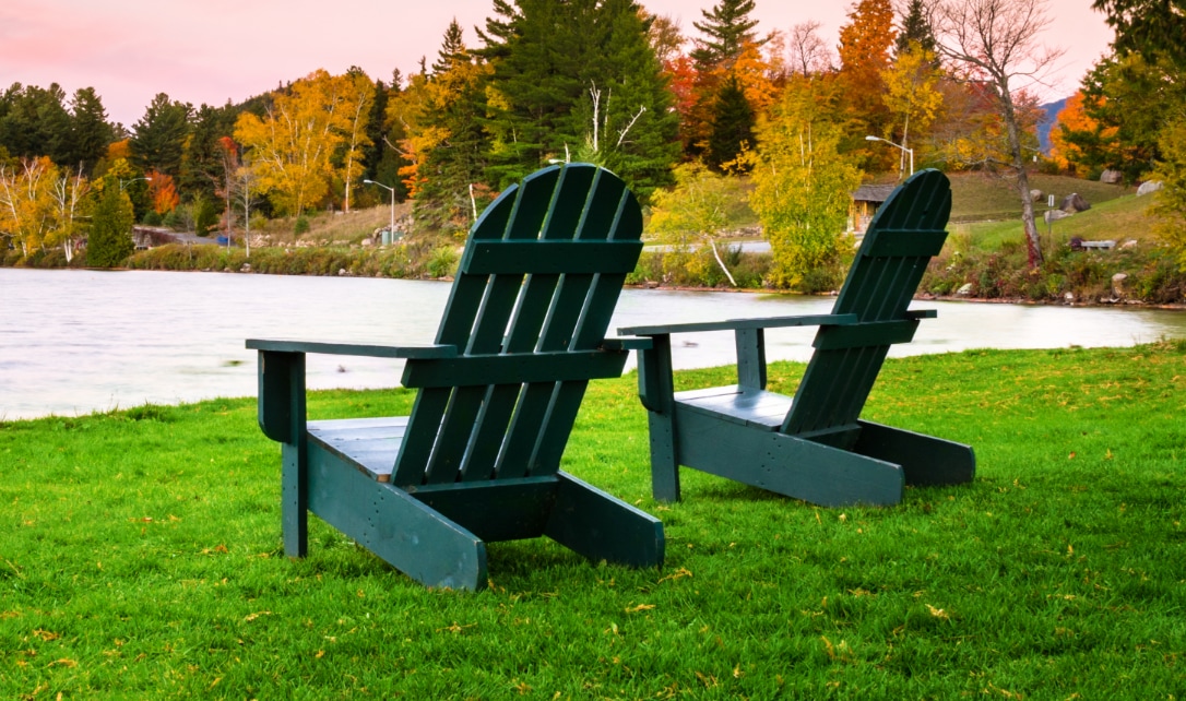 Two Adirondack chairs on a grassy lawn overlooking a lake with autumn trees in the background.