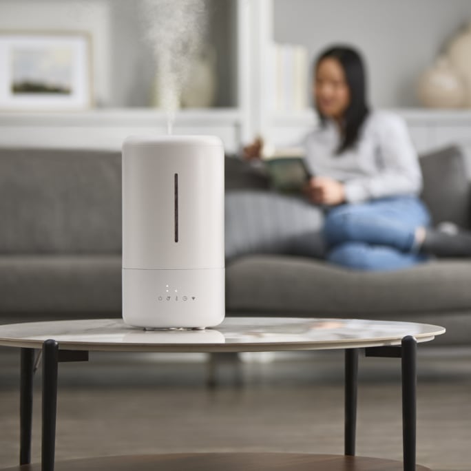 A humidifier stands on a coffee table with a person sitting on a couch in the background.