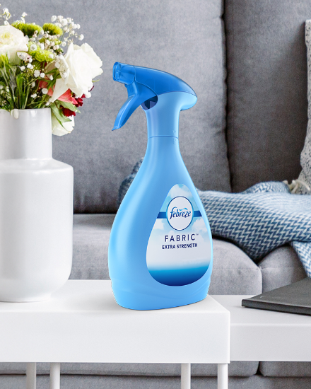 Blue spray bottle of fabric freshener on a white table, with a sofa and decorative pillows in the background. Flowers are also visible in a vase.