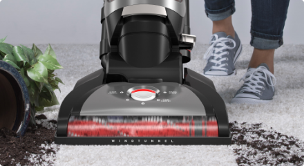 Upright vacuum cleaner being used to clean mud from a fallen potted plant on a white carpeted floor.