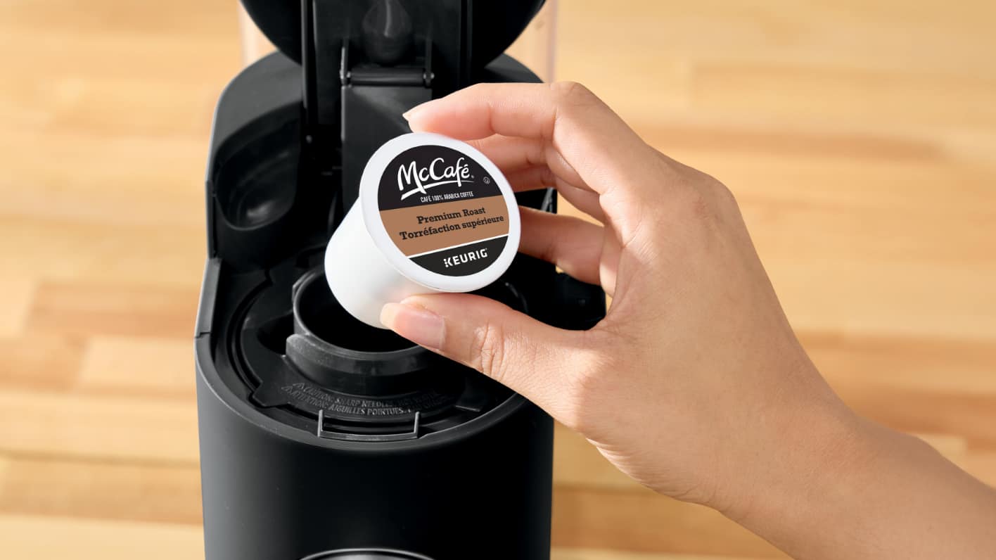 Hand placing a coffee pod into a coffee maker. Text reads "Coffee Pods" and includes a "Shop Now" button.