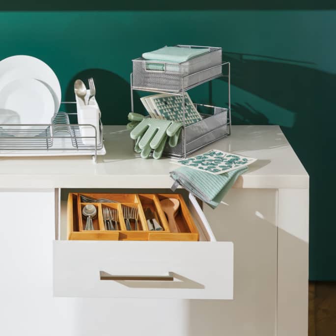 Organized kitchen drawer with neatly arranged utensils and a countertop with dishes and a rack of cleaning cloths.