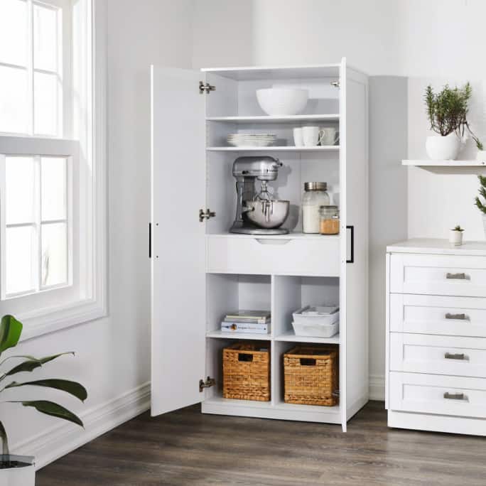 White pantry cabinet in a bright kitchen, with open doors revealing shelves of appliances, containers, dishes, and storage baskets.