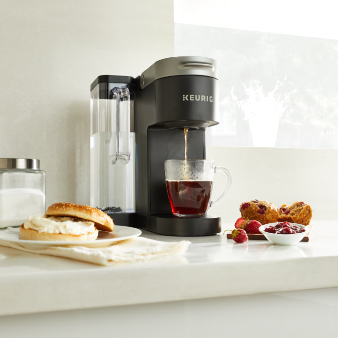 Single serve coffee maker on a white kitchen counter next to plates with a bagel, muffins, strawberries, and a bowl of jam.