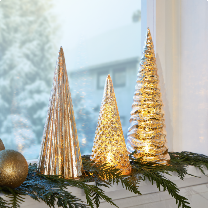 Three decorative, lit gold trees on a windowsill with greenery and ornaments.