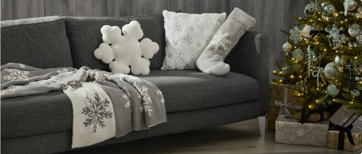 A gray couch with snowflake-themed pillows and blankets next to a decorated Christmas tree with gifts underneath.