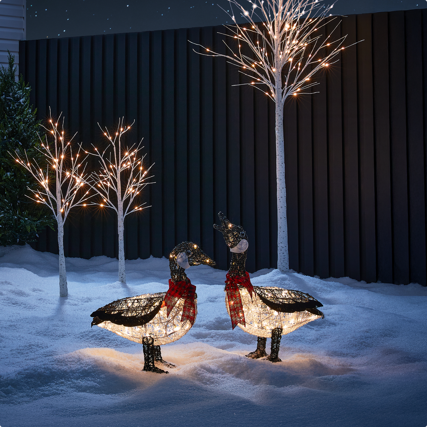 Two illuminated CANVAS Birch Trees and two CANVAS LED Canadian Cabin Geese decorations displayed in a snowy front lawn at night.