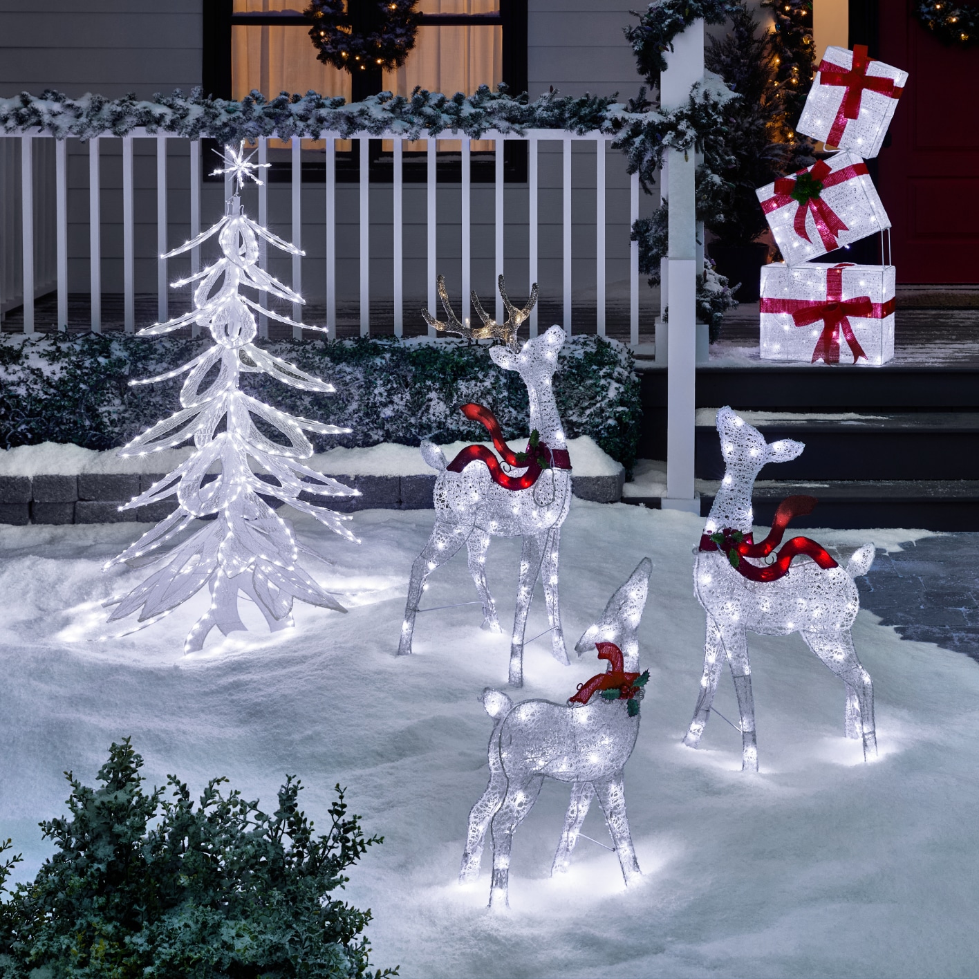 A CANVAS Arctic Gift Box set decoration, three CANVAS Arctic Deer, and a CANVAS Arctic Twinkling Tree displayed in a snowy front lawn at night.