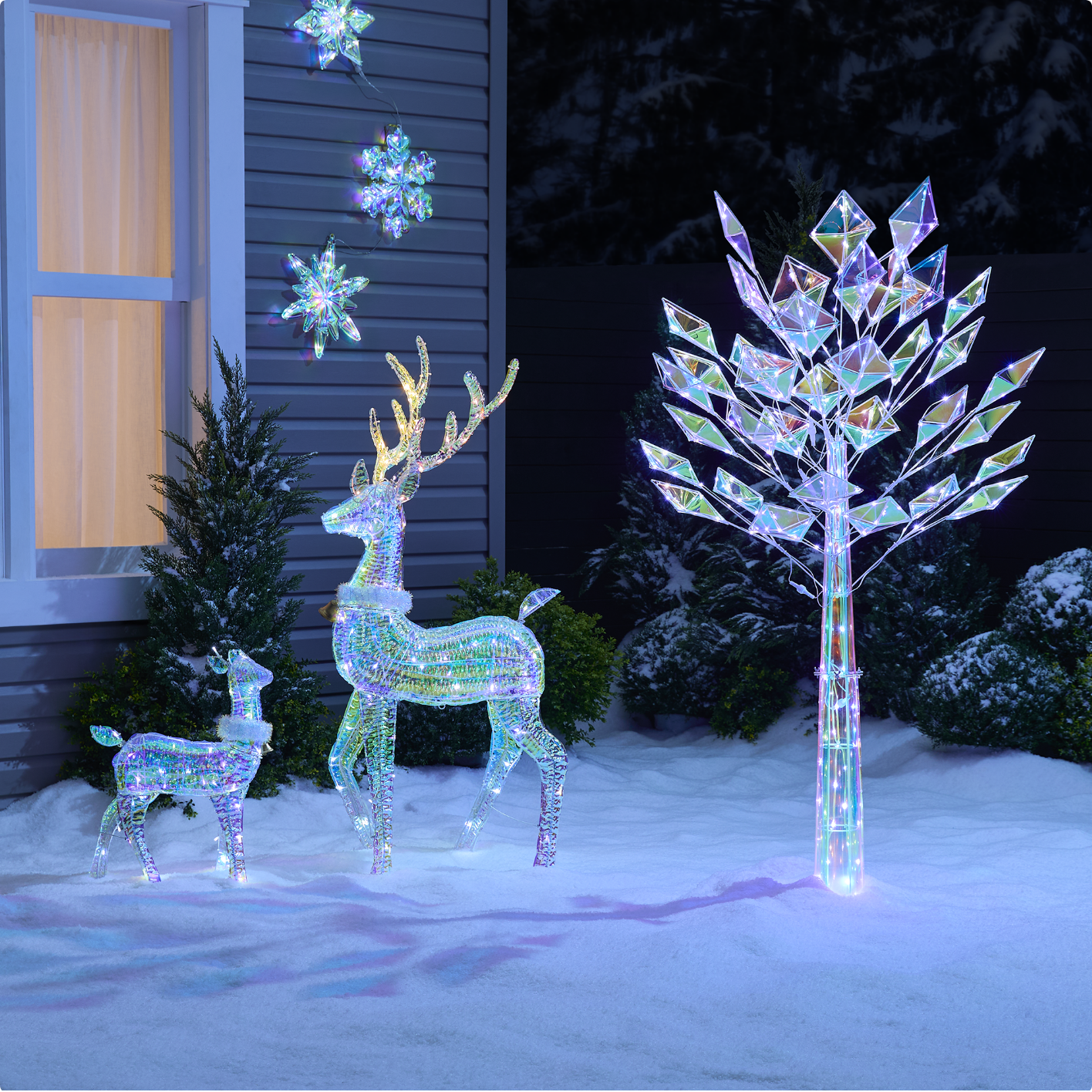 A CANVAS Glisten Twinkle Tree, CANVAS Glisten Deer & Fawn decorations, and CANVAS Glisten Hanging Snowflakes displayed in a snowy front lawn at night.