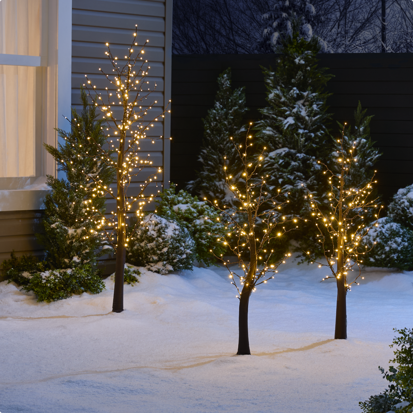Three illuminated CANVAS Blossom Twinkle Trees displayed in a snowy front lawn at night.