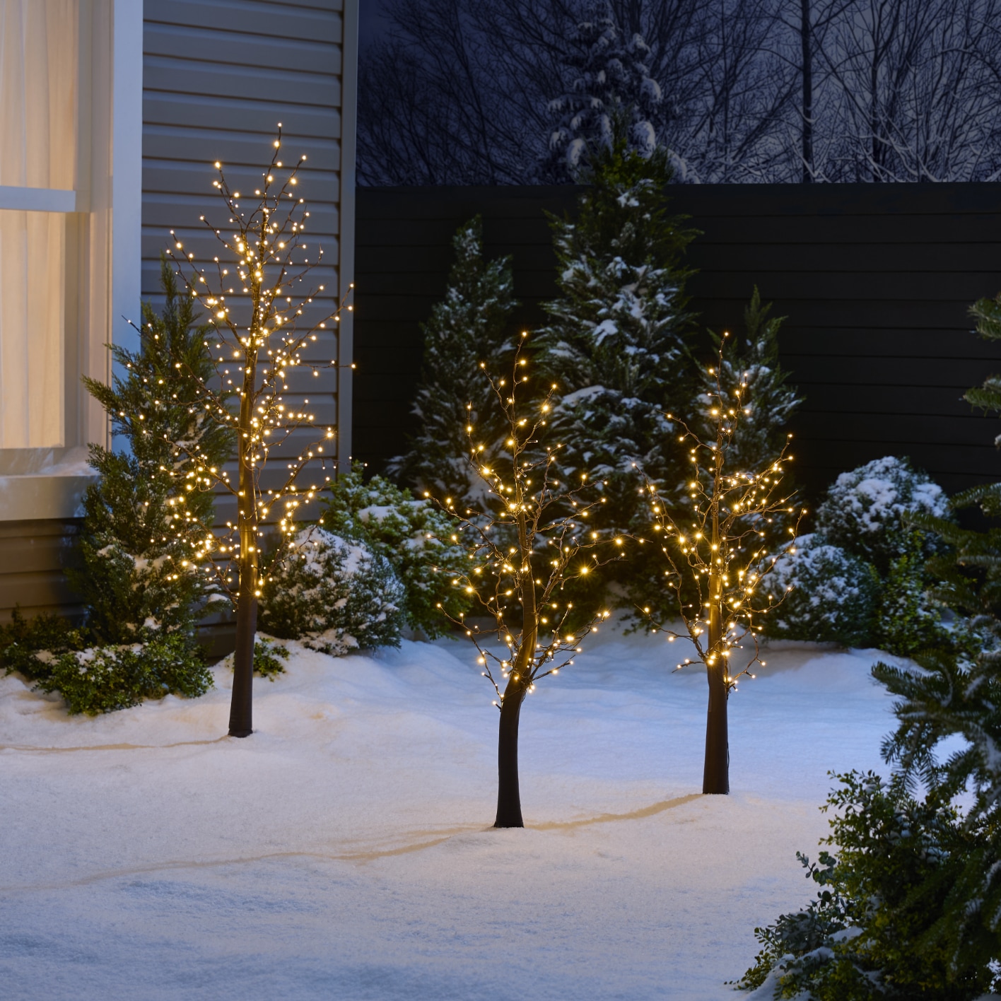 Small trees adorned with twinkling lights create a festive atmosphere in a snow-covered yard