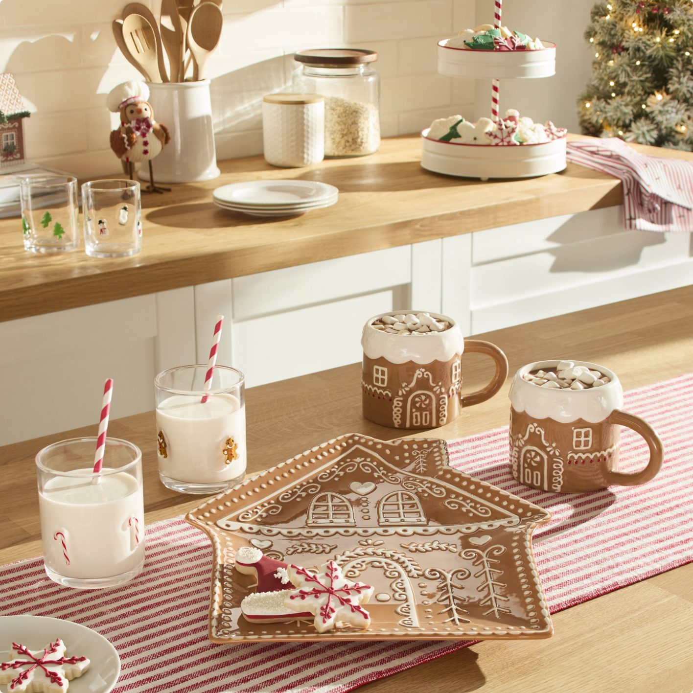 A kitchen counter with gingerbread-themed mugs, milk glasses with striped straws, and a gingerbread house plate on a red-striped runner.