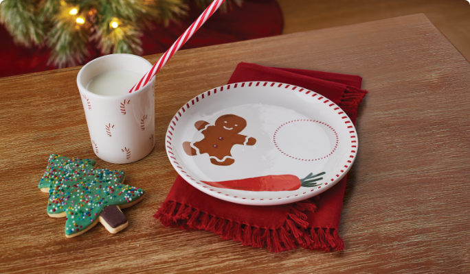 Plate with a gingerbread design sits on a table next to a Christmas tree cookie, and a candy cane-themed mug.