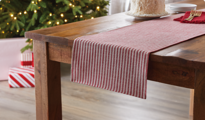 A wooden table with a red and white striped runner, a cake, and a Christmas tree with lights in the background.
