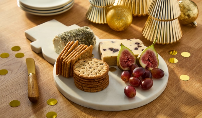 Plateau de fromages et de crackers accompagné de figues et de raisins sur une table, entouré de miniatures dorées décorant le sapin de Noël.