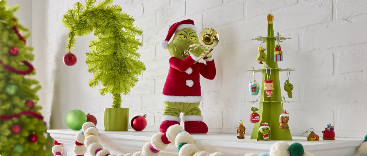 Un arbre Crumpit For Living de 4,5 pi et divers articles de décoration sur le thème du Grincheux disposés dans une pièce lumineuse.  Un assortiment de mini ornements incassables Le Grincheux, de décorations en résine du Grincheux sur une échelle et de décorations Le Grincheux en verre soufflé sur un arbre Crumpit For Living inspiré du Grincheux.